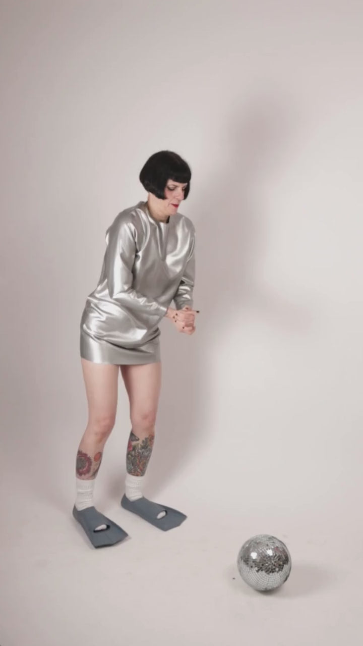 Anja von Abels is wearing a silver latex sweatshirt dress and fins. In this video clip she is kicking a disco mirror ball almost like a soccer pro.