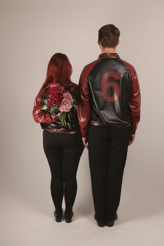 Rear shot of Sarah and Paul standing attention in their matching black and bordeaux-red latex college jackets. Sarah is very obviously hiding a bouquet of flowers behind her back.