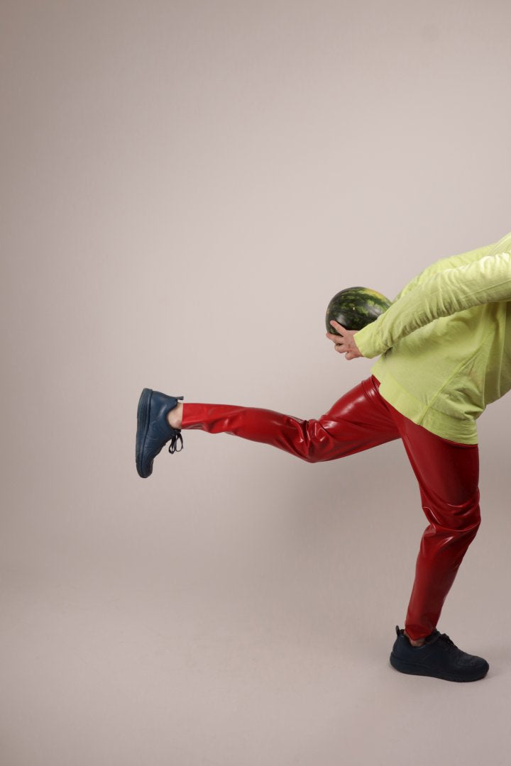 Fabian in glossy loose red latex leggings in a ministry of silly walks move and carrying a watermelon