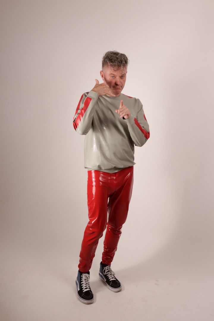 Fabian doing 'i'll call you' sign language  towards the camera in his grey latex sweatshirt with red stripes on the arms, and red latex leggings and SK8 hi top Vans.