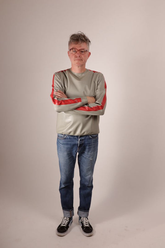 Front view of Fabian in Vans, blue jeans and his grey latex sweatshirt with red stripes on the arms. Arms crossed.