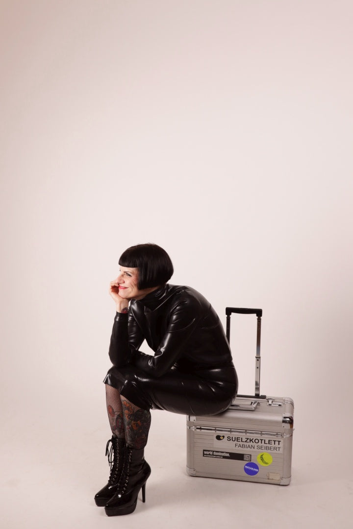 Our model Anja grinning and sitting on an aluminum flight case in our black latex pencil skirt and matching latex loose turtleneck