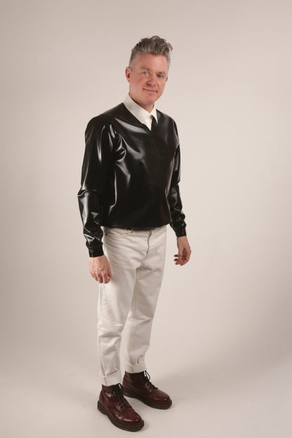 Fabian smiling into the camera in his black V-neck latex sweater over a white dress shirt and with white skinny jeans and cherry red Dr. Martens boots.