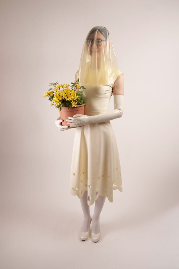 Hani wearing our TARZA & JANE lacey wedding dress. The sleeveless silhouette with wide shoulders and a t-shirt neckline looks just lovely with the midi skirt and its lace pattern hem. Hani wears it with a transparent latex veil and long white latex opera gloves. Quite cute is the pot with yellow flowers instead of the bouquet.