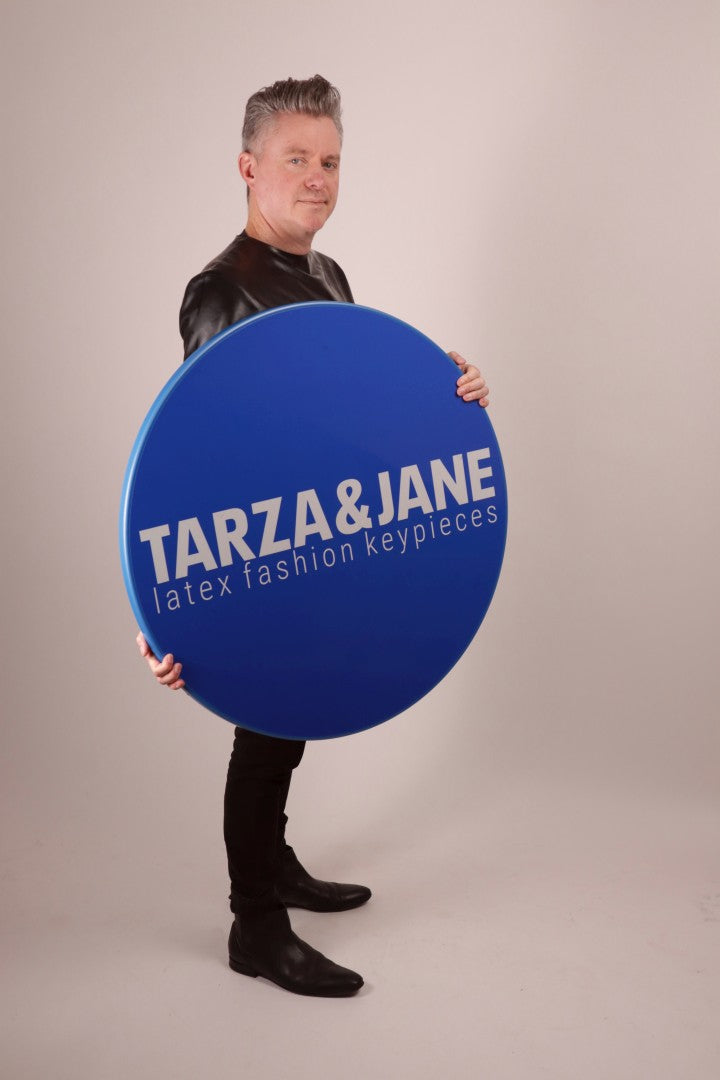 Fabian holding a huge round blue TARZA & JANE traffic sign. The sign is so big it almost completely covers Fabians latex sweater.