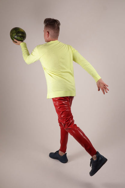 Fabian in glossy loose red latex leggings, a neon yellow longsleeve and blue camper sneakers doing a bowling move with a watermelon